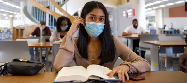 student sitting at library table with an open book wearing a mask