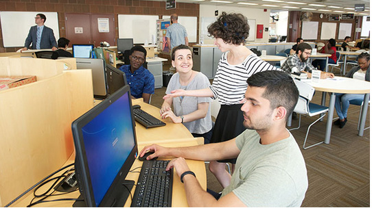 two students sitting at computers talking to a tutor who is standing nearby