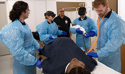 Paramedic students standing near a manikin on a table; one student is holding a manikin baby