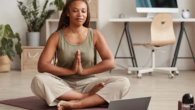 woman in a meditation pose sitting on a yoga mat on the floor in front of a laptop computer